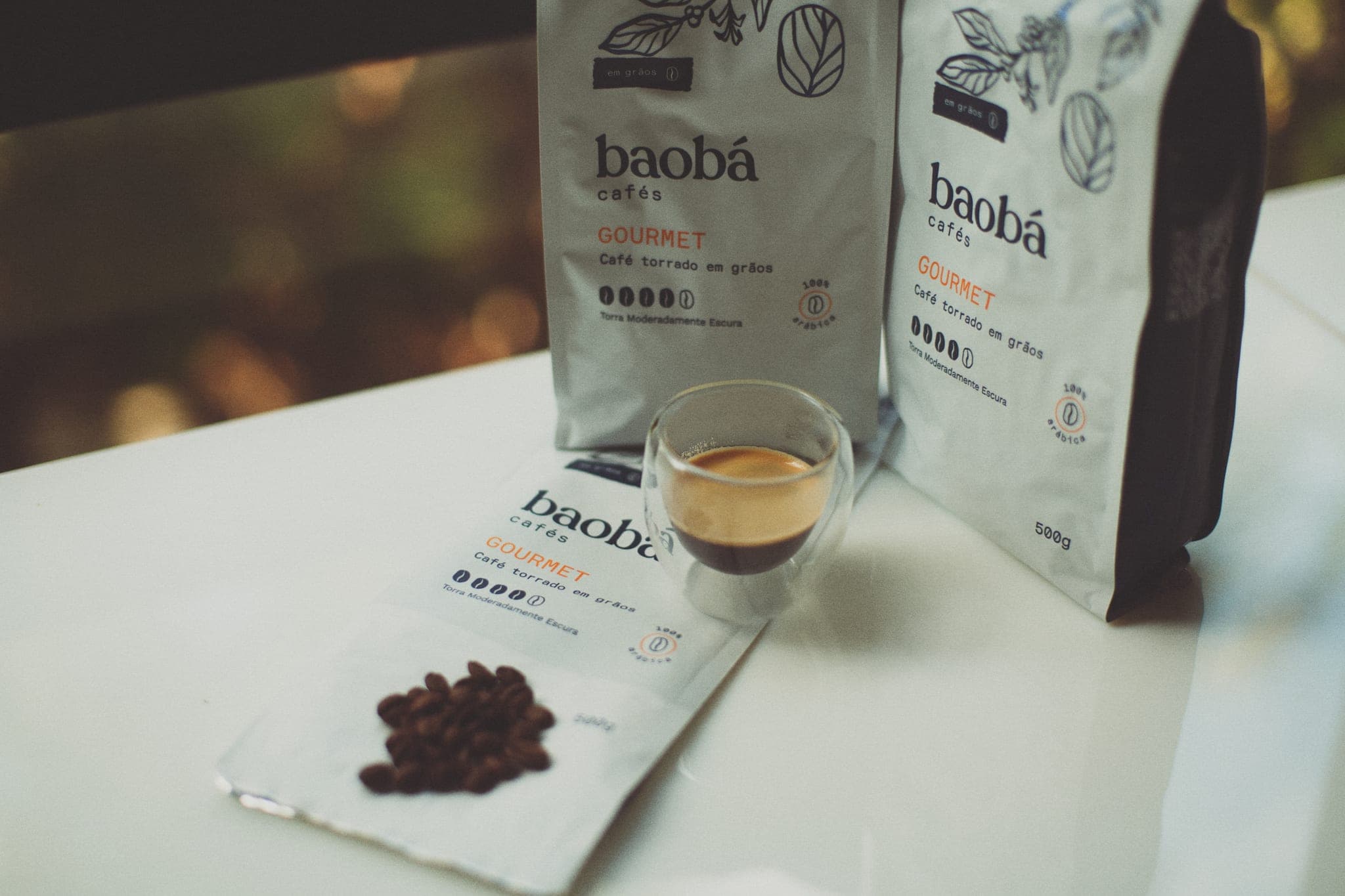 Dois pacotes de 500 g do Baobá Gourmet em grãos ao lado de uma xícara de vidro de parede dupla com café, e um terceiro pacote aberto com grãos torrados sobre uma mesa clara
