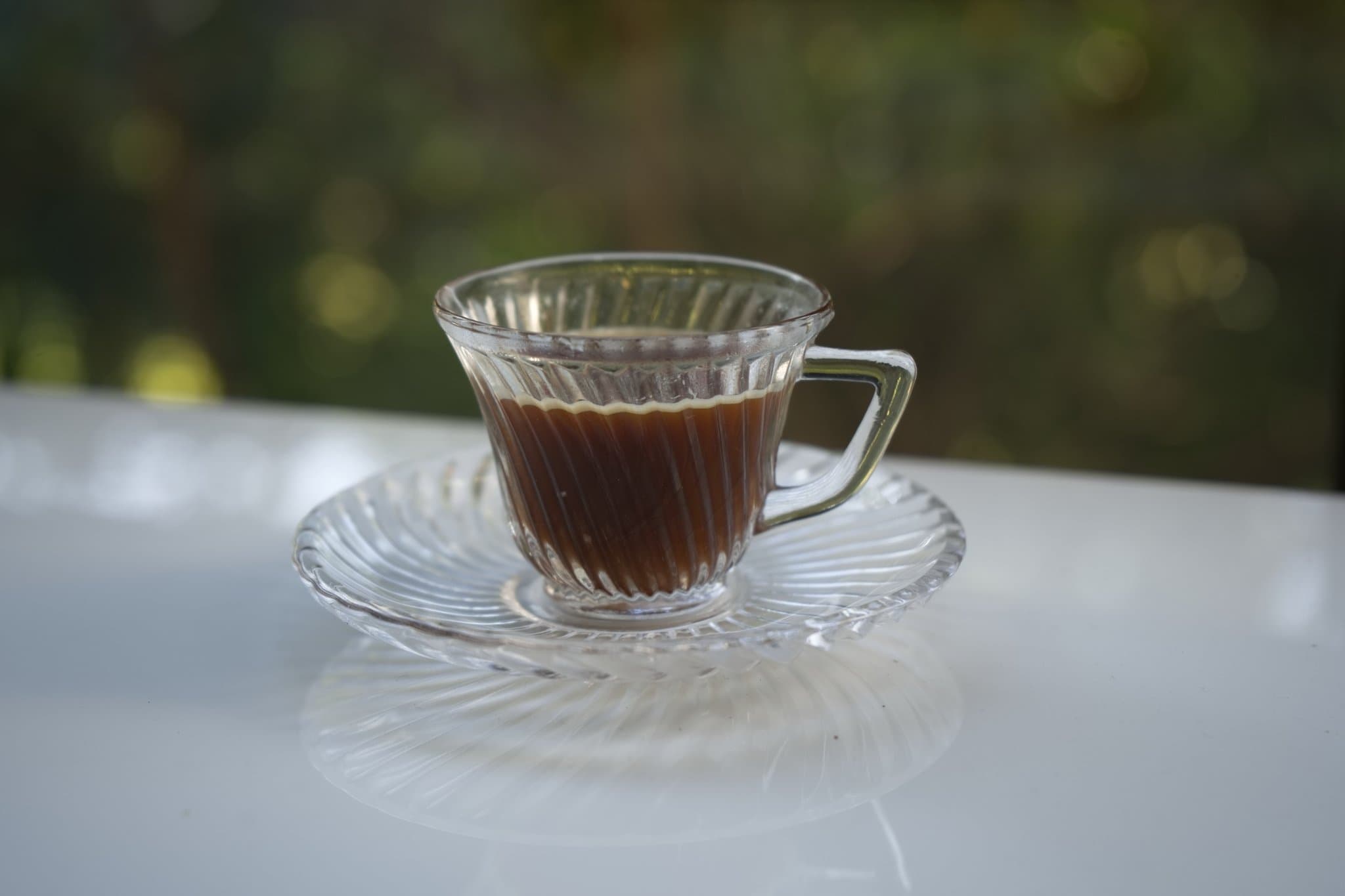 Café Gato-Mourisco servido em xícara de vidro sobre pires, com vegetação ao fundo