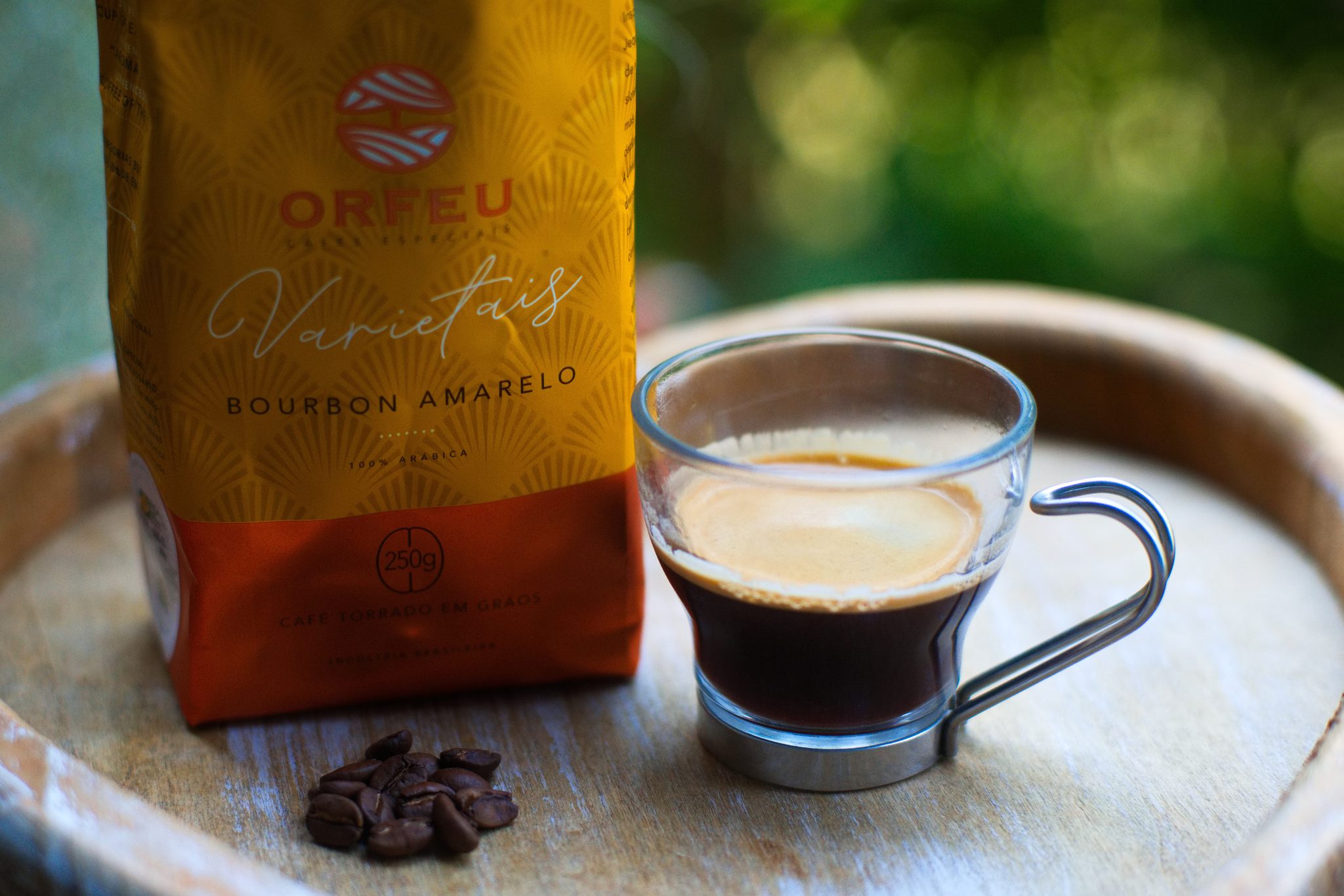 Pacote do Orfeu Varietais Bourbon Amarelo ao lado de uma xícara de vidro com café e alguns grãos sobre a bandeja de madeira